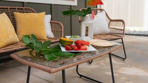 Un sofá de mimbre con cojines amarillos, una mesa de vidrio con frutas y una planta, y un jarrón blanco con una flor.