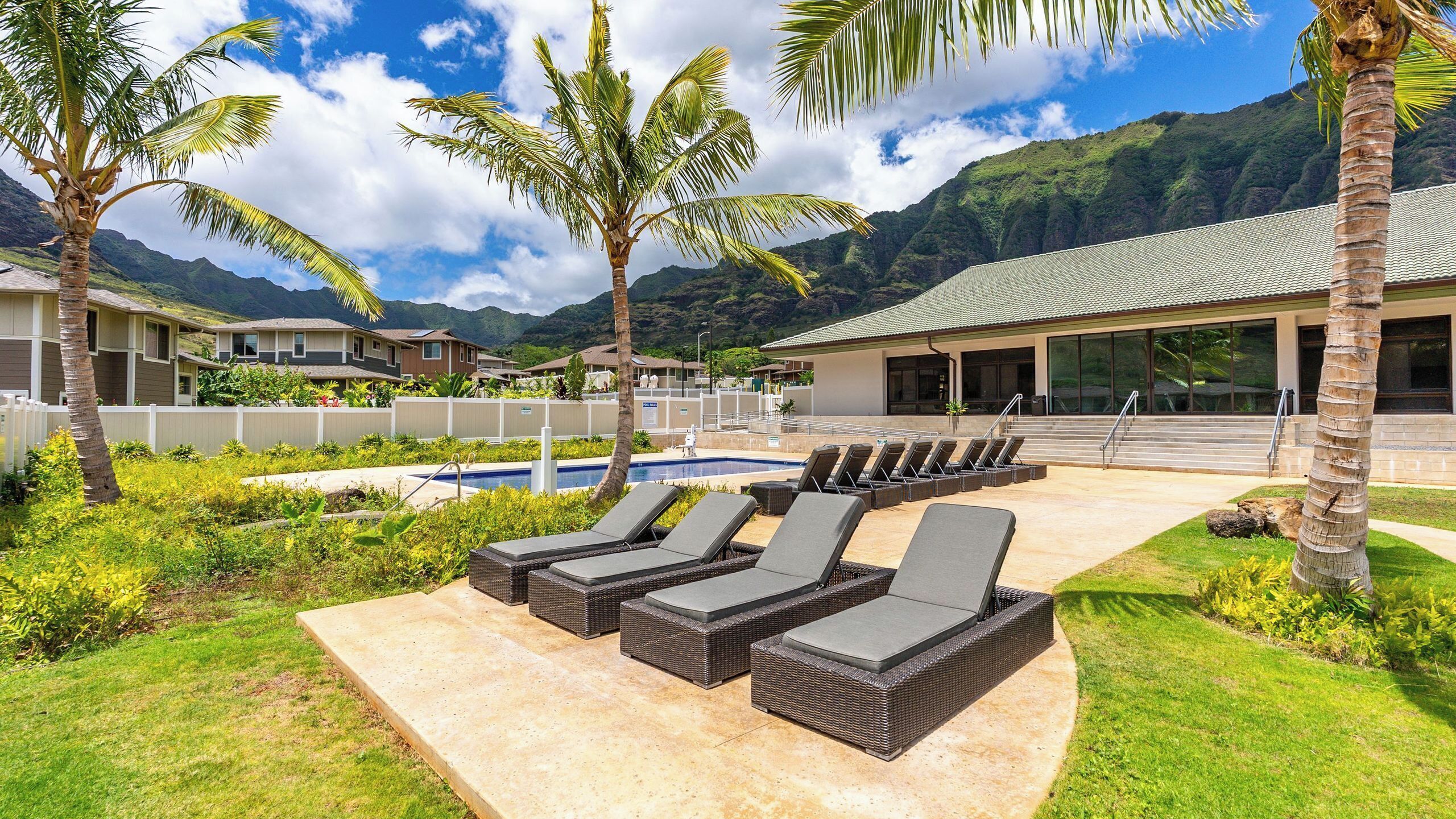 Un área de piscina con sillones de descanso, palmeras y un edificio con techo de tejas.