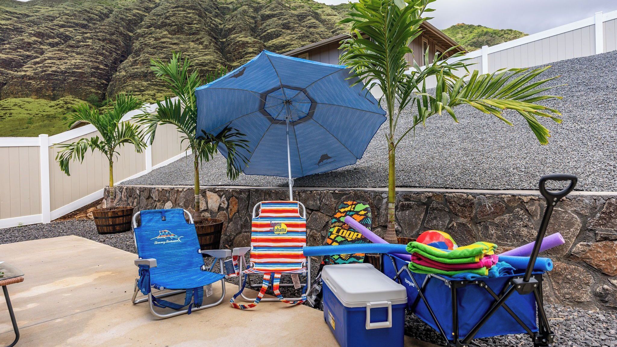 Un área de patio con un paraguas azul, una silla de playa a rayas, un enfriador y una silla plegable.