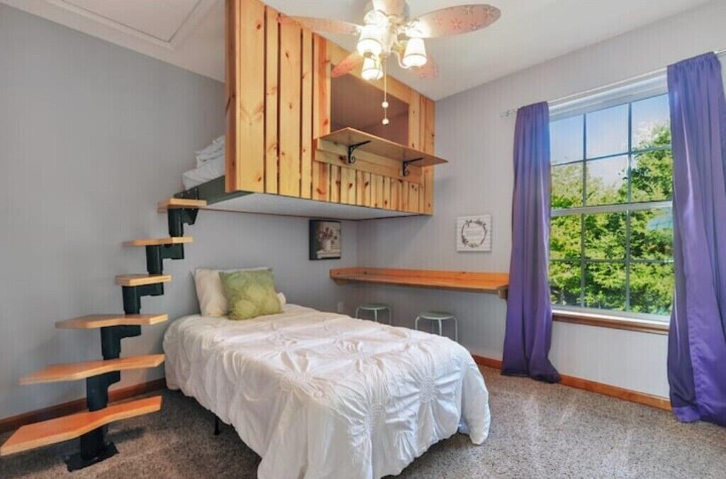 Ein Schlafzimmer mit Bett, einer Holztreppe, einem Deckenventilator, einem Fenster mit Vorhängen und Blick ins Grüne.