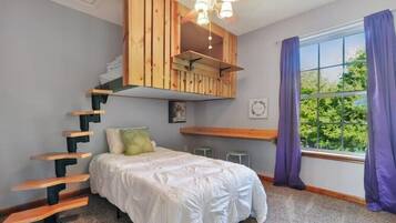 Ein Schlafzimmer mit Bett, einer Holztreppe, einem Deckenventilator, einem Fenster mit Vorhängen und Blick ins Grüne.