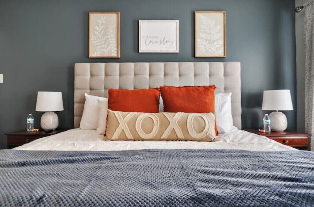 Un letto rifatto con una coperta a righe grigie e bianche, due abat-jour e un poggiatesta con un cuscino 'XOXO'.