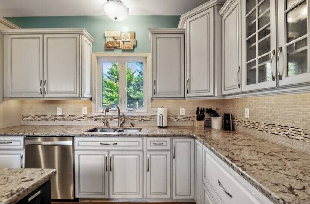 A kitchen with white cabinets, a granite countertop, a stainless steel dishwasher, and a window with a view of trees.