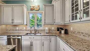 A kitchen with white cabinets, a granite countertop, a stainless steel dishwasher, and a window with a view of trees.