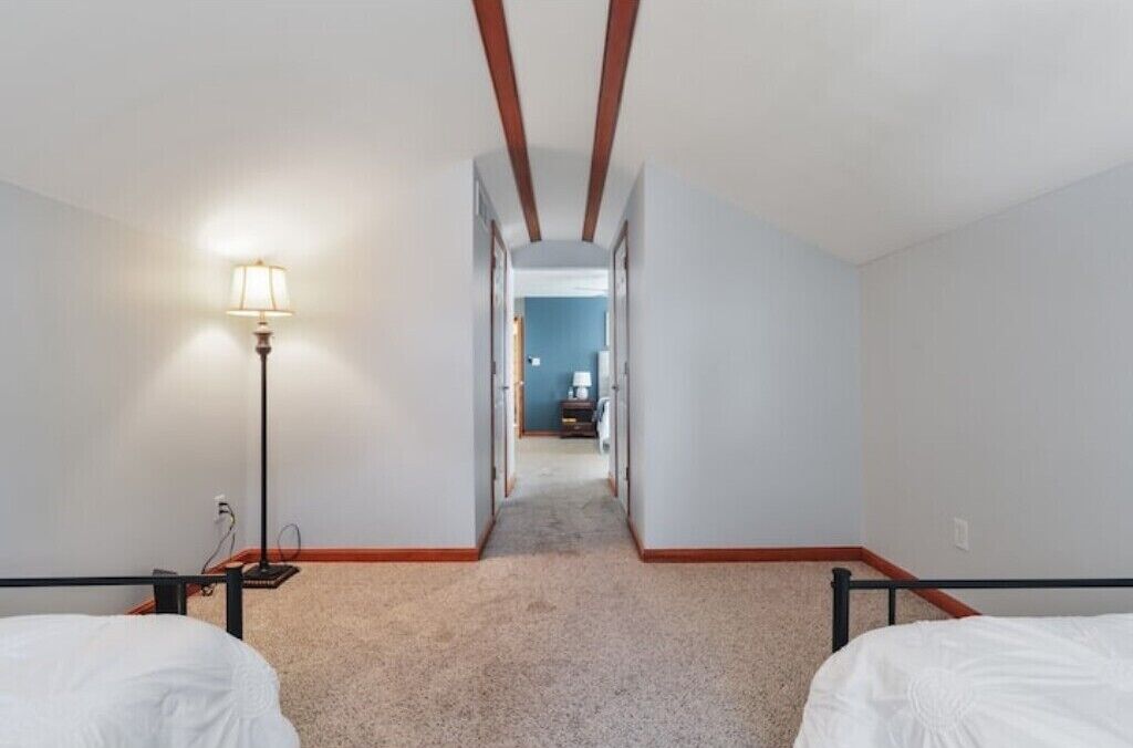 Habitación de hotel con piso alfombrado, cama, mesita de noche y lámpara. Se ve un pasillo que lleva a otra habitación.