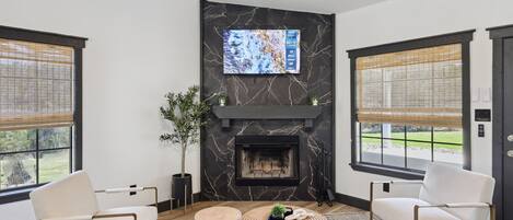 Una sala moderna con chimenea, dos sillones blancos, una mesa de centro y un televisor montado en una pared de mármol.