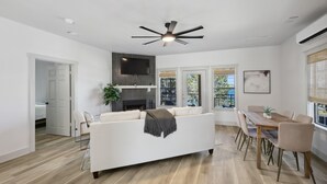 A modern living room with a white sofa, dining table, and chairs. There is a fireplace, a ceiling fan, and large windows with blinds. - The Retreat 20: Lake Daze (Graford)
