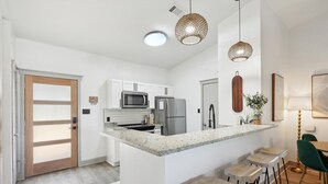 A modern kitchen with white cabinets, a granite countertop, and a central island with bar stools. There are pendant lights above the island and a refrigerator to the right.