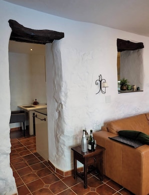 A living room with a brown leather sofa, a small wooden side table with bottles, a tiled floor, and a kitchen area in the background. - La Casa Antigua, Canillas de Albaida (Canillas de Albaida)
