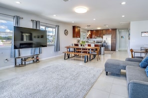 Una sala moderna con un televisor de pantalla plana, comedor con mesa y sillas de madera, un sofá gris y una cocina con armarios de madera oscura y electrodomésticos de acero inoxidable.