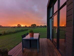 A wooden deck with a table and chairs, overlooking a grassy field and a sunset.