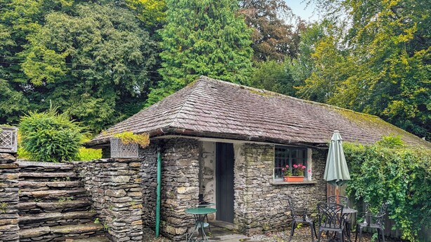 Une petite maison en pierre avec un toit en tuiles, un petit porche et un jardin aménagé avec des chaises et une table.