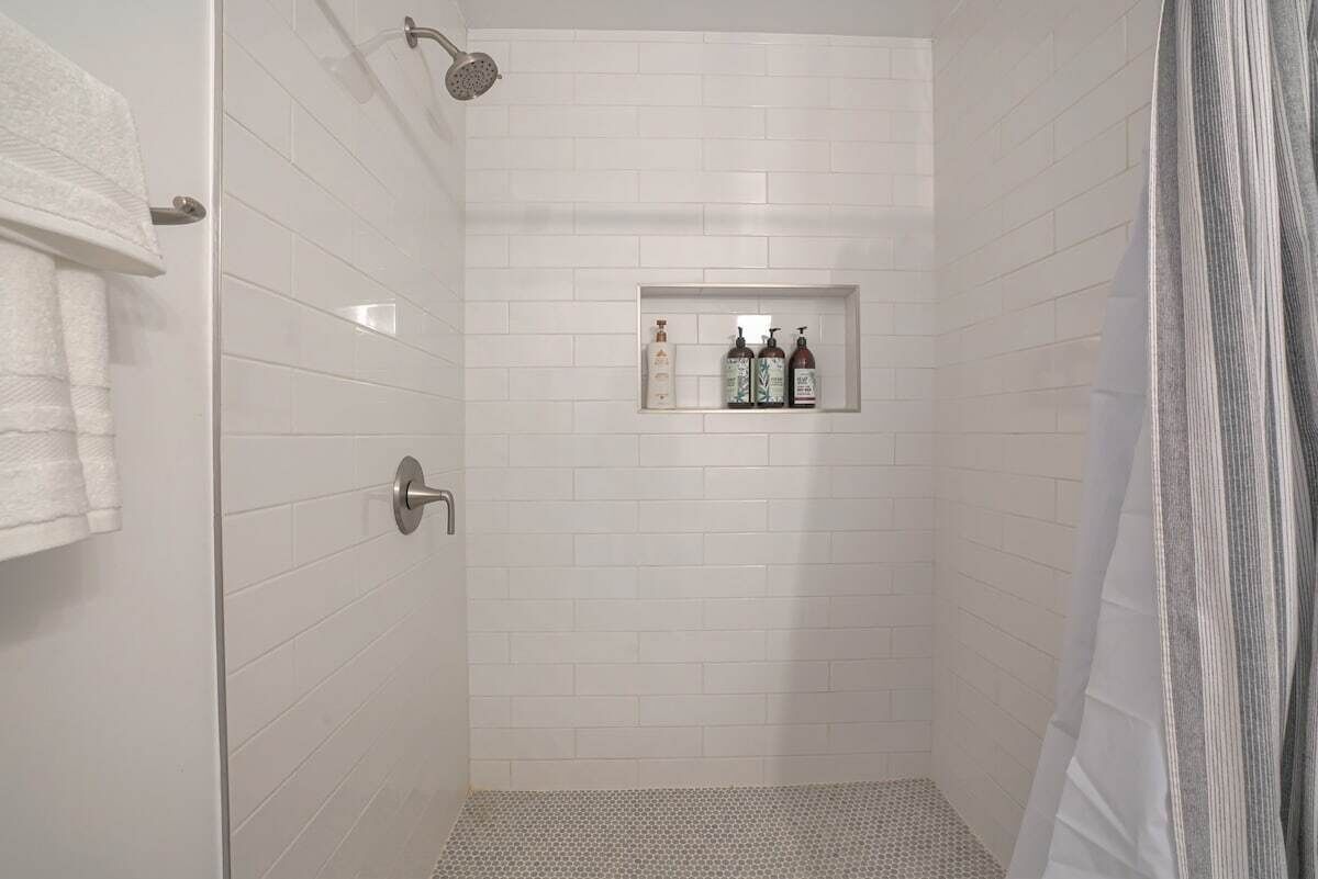 A shower with white tiled walls, a showerhead, and a shelf with toiletries.