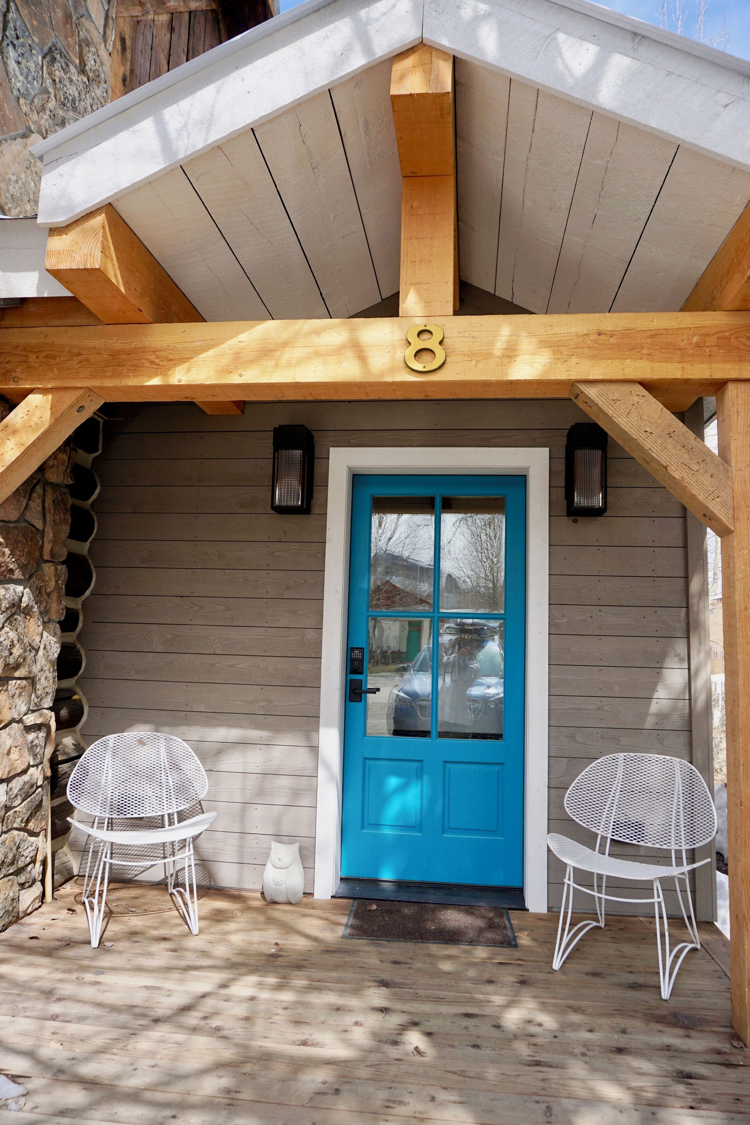 A wooden cabin with a blue door, two white metal chairs, and a stone wall.