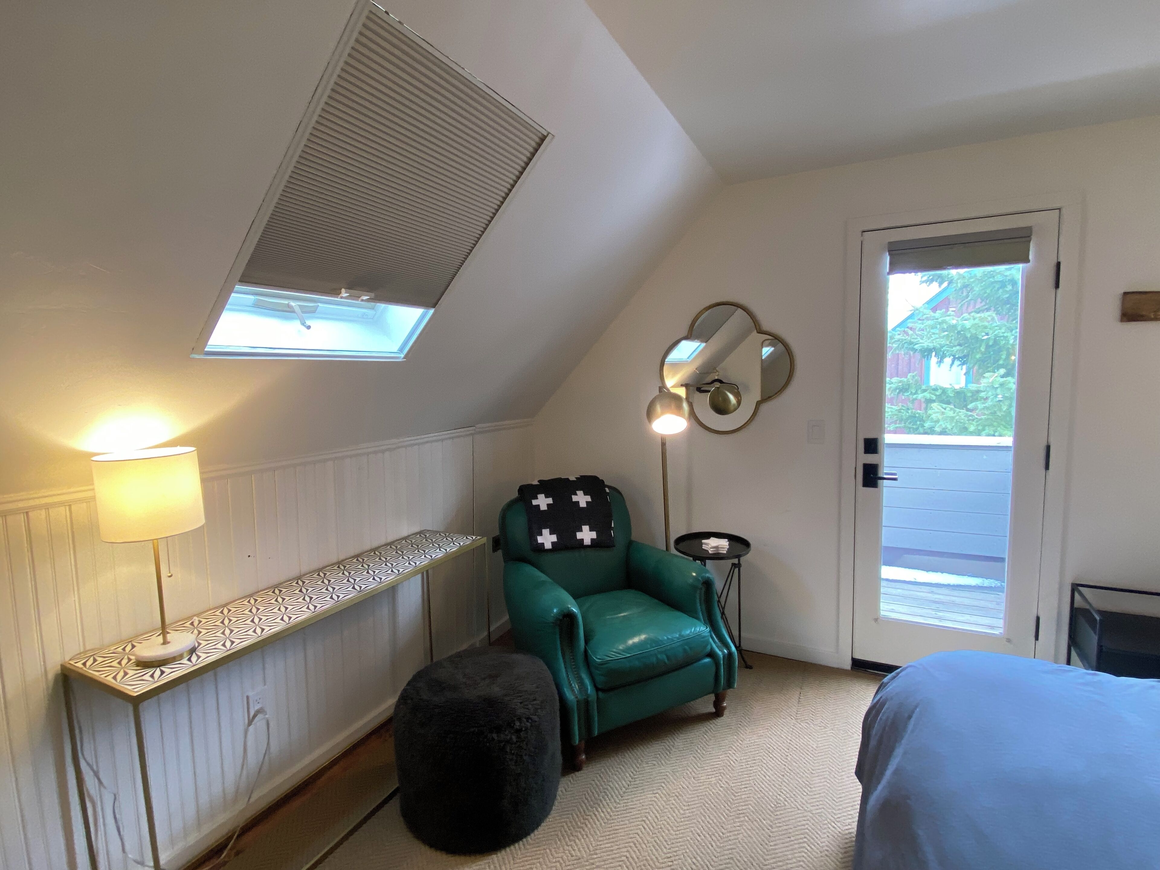 Una habitación con techo inclinado, una ventana con persianas, un sillón verde, un banquillo redondo pequeño, una mesita de noche con una lámpara y una puerta que da al exterior.