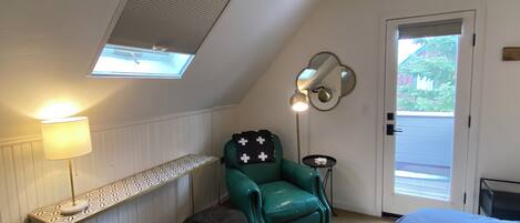 Una habitación con techo inclinado, una ventana con persianas, un sillón verde, un banquillo redondo pequeño, una mesita de noche con una lámpara y una puerta que da al exterior.