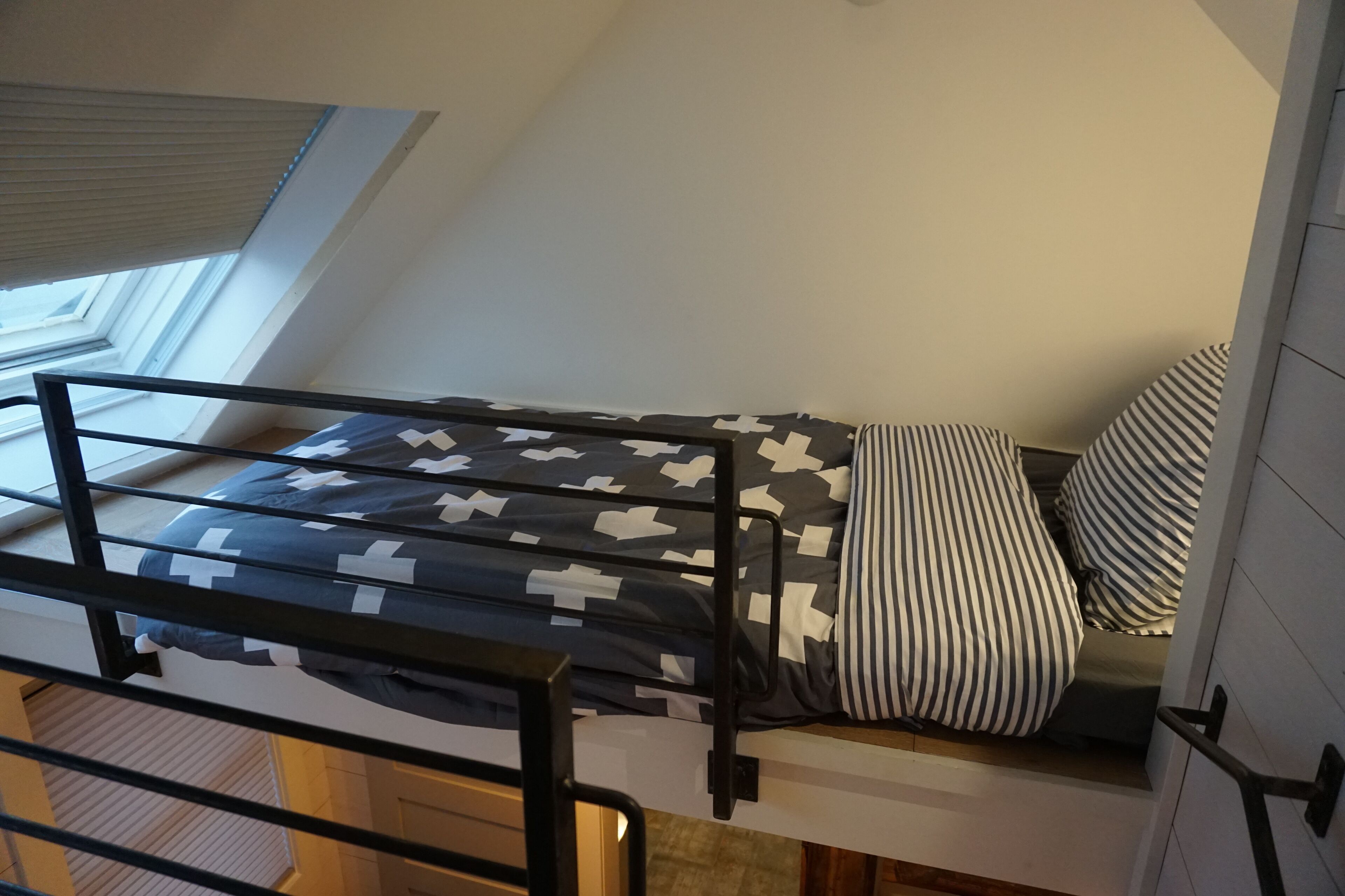 Una cama individual con edredón estampado y almohada a rayas, una ventana con persianas y vistas al exterior.