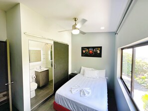 Une chambre d’hôtel avec un lit, une salle de bain, un ventilateur de plafond et une fenêtre donnant sur un beau paysage.
