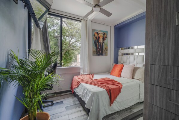 Une chambre à coucher avec un lit, un ventilateur de plafond, une fenêtre avec des rideaux, un tableau représentant un éléphant et une plante en pot.