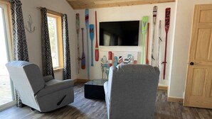A living room with two grey armchairs, a flat-screen TV, and a wall decorated with various paddles. - Anna Bananas Beachfront Bungalow (Hazelhurst)