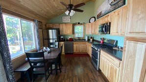 A kitchen with wooden cabinets, a dining table with chairs, a ceiling fan, and a window with curtains. - Anna Bananas Beachfront Bungalow (Hazelhurst)