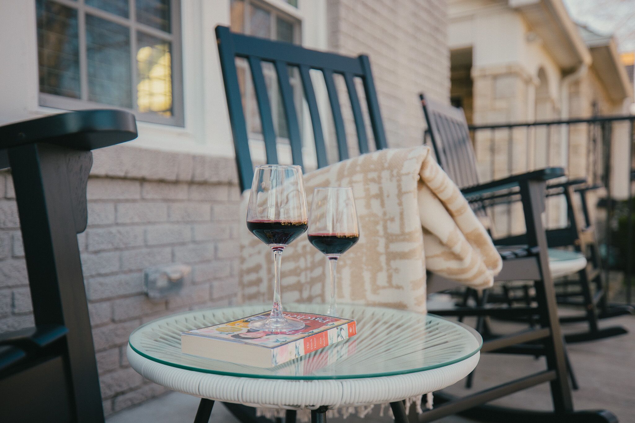 Una mesa de vidrio con dos copas de vino y un libro, sobre un balcón con una silla y una manta.