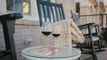 Una mesa de vidrio con dos copas de vino y un libro, sobre un balcón con una silla y una manta.