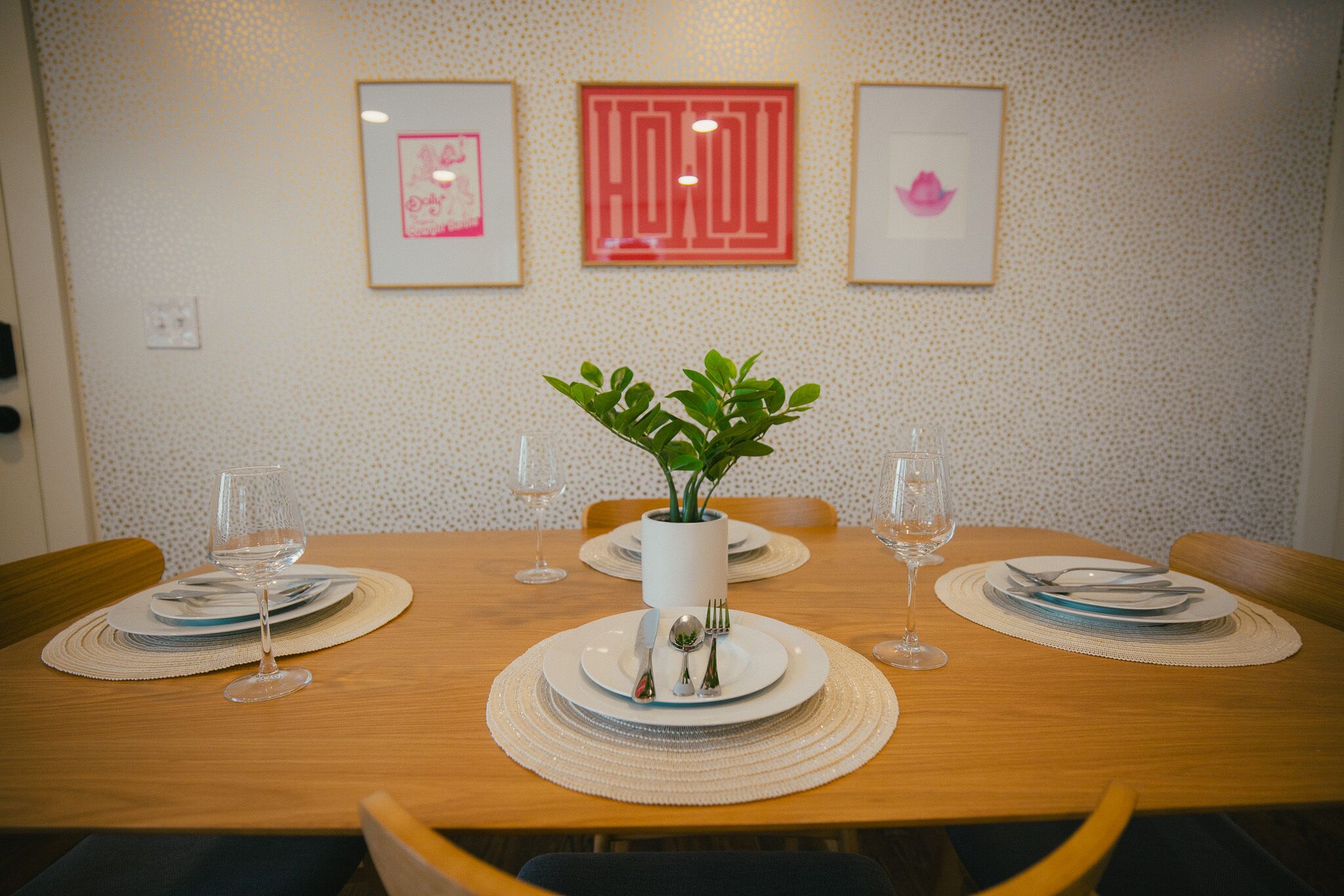 Una mesa de comedor dispuesta con platos, cubiertos y vasos, con una planta en maceta en el centro.