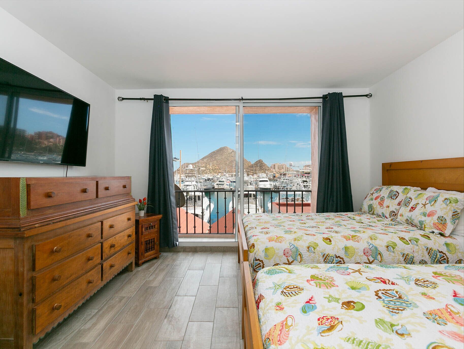 A bedroom with a bed, dresser, and a TV. It has a balcony with a view of boats and a mountain.