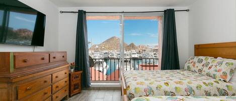 A bedroom with a bed, dresser, and a TV. It has a balcony with a view of boats and a mountain.