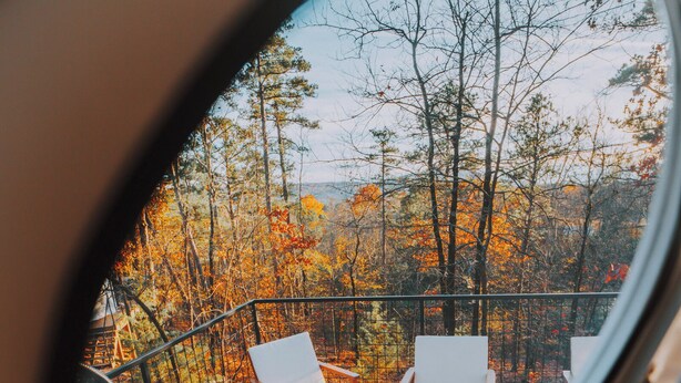 A circular window view of a deck with four white lounge chairs and a small table, overlooking a forested area with autumn foliage. - Mockingbird Geodesic Dome at Starlight Haven Resort (Hot Springs)