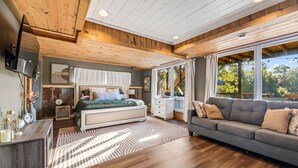Un dormitorio moderno con techo de madera, una cama grande con colcha verde, un sofá gris y un amplio ventanal con vistas al exterior.