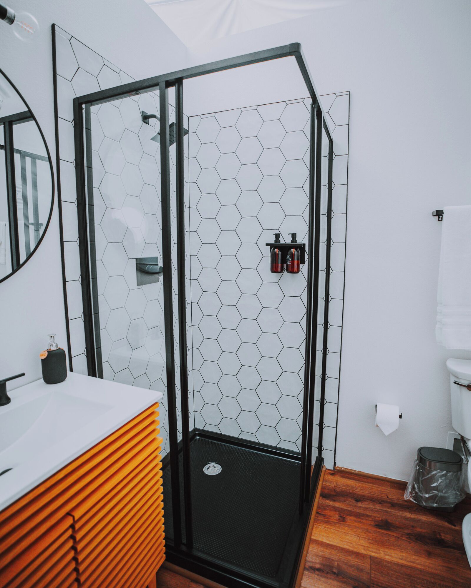 A modern bathroom with a glass shower enclosure, white hexagonal tiles, a wooden vanity, and a mirror.