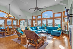A spacious living room with a large blue sectional sofa, wicker chairs, and a dining area with wooden chairs and a glass table.