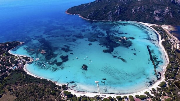 Eine Luftaufnahme einer Küstenregion mit klarem türkisblauem Wasser, einem Sandstrand und einem gebirgigen Hintergrund.