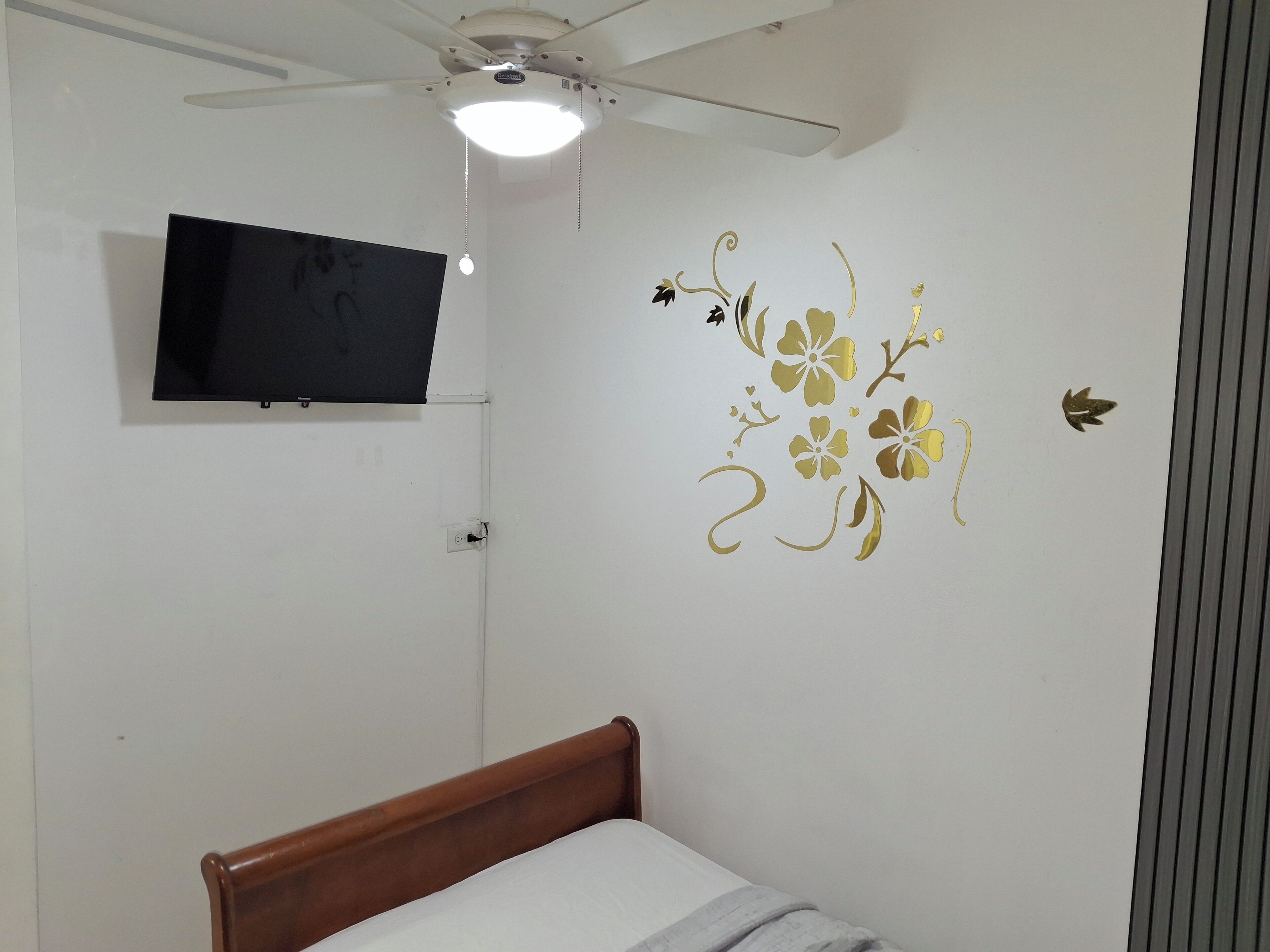 Quarto com cama, ventoinha no teto, TV fixada na parede e adesivos decorativos.