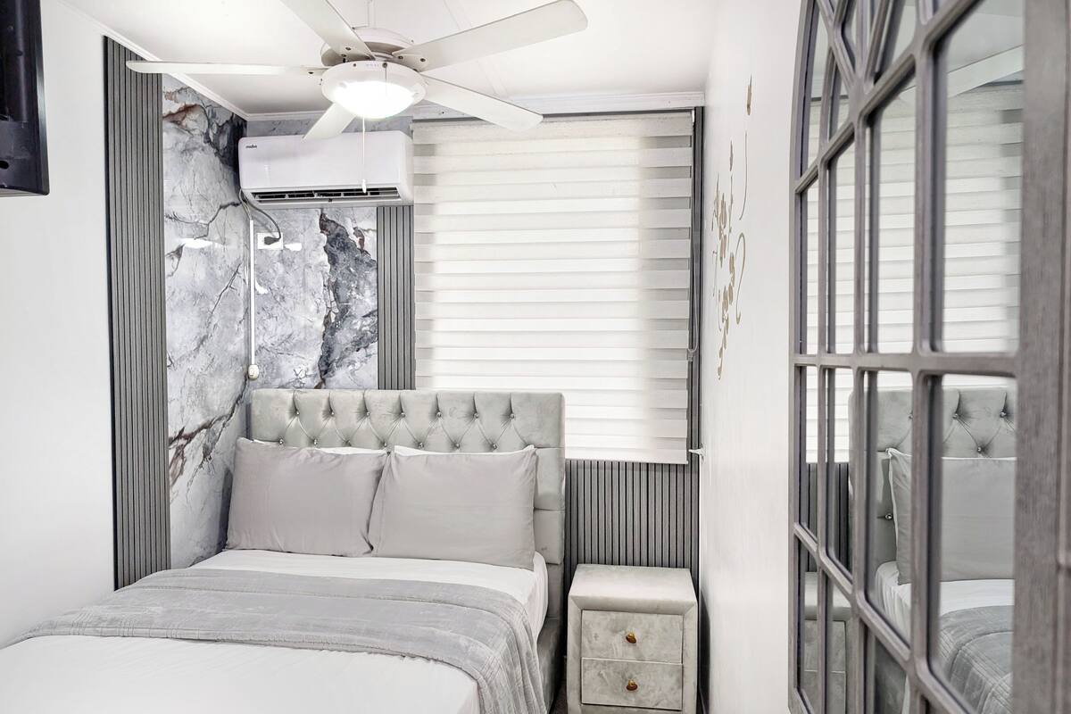 Quarto moderno com cabeceira estofada, mesa de cabeceira com gavetas e ventilador de teto.