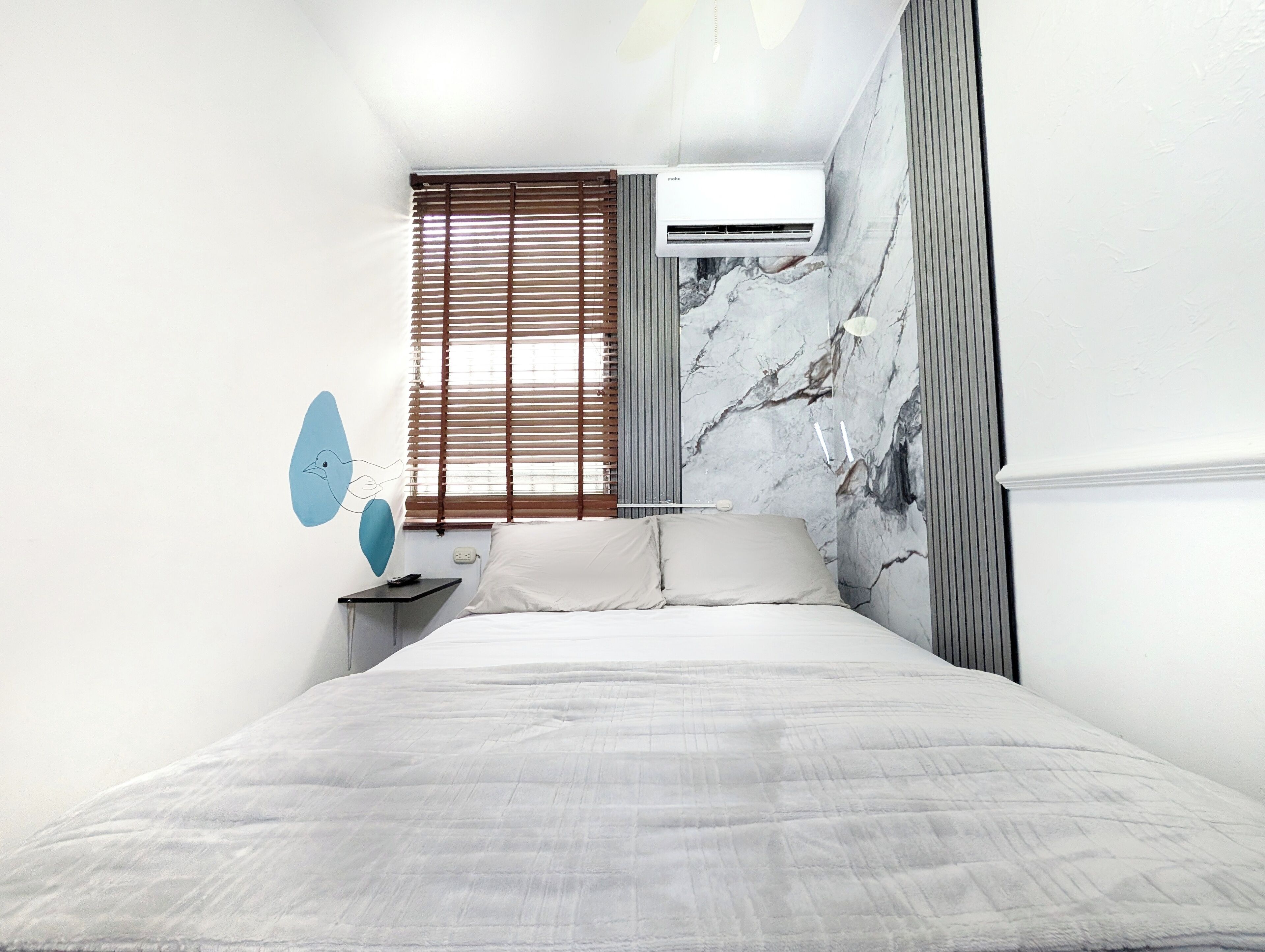 Ein Schlafzimmer mit einem Bett, einem Nachttisch, einer Klimaanlage und einem Fenster mit Jalousien.