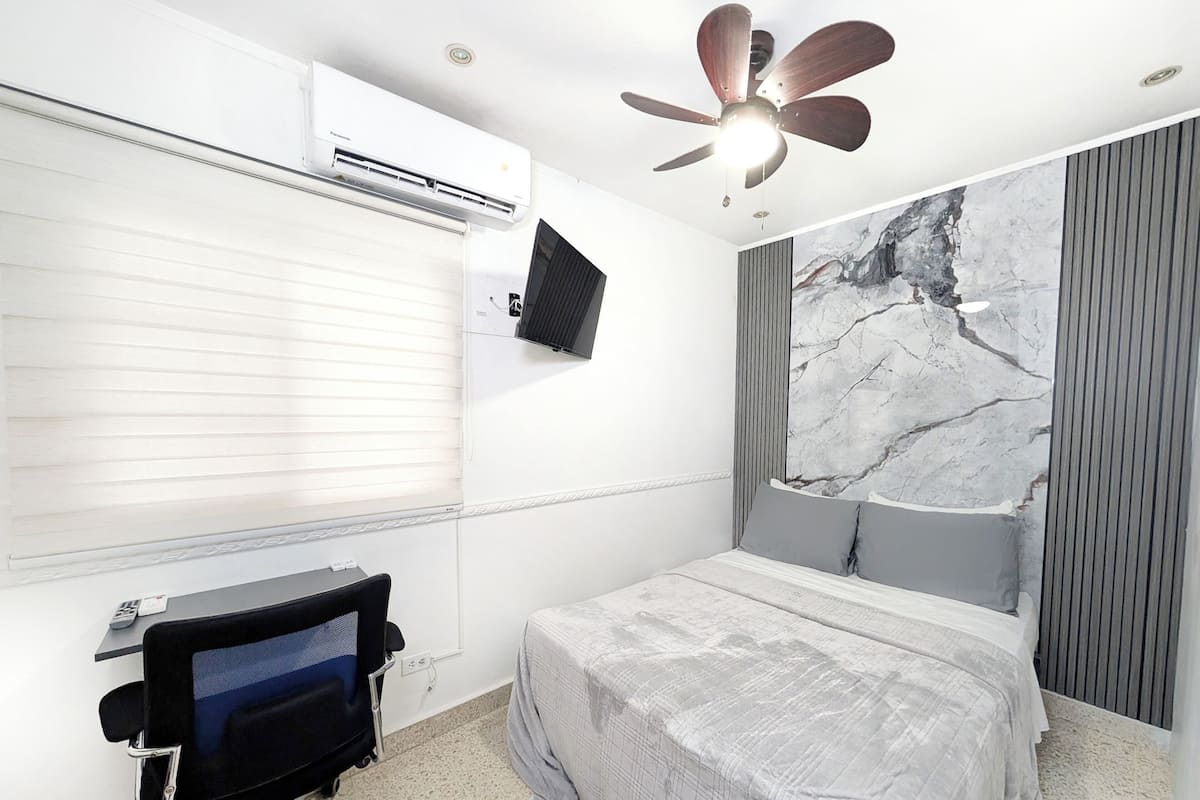 Um quarto com uma cama, uma escrivaninha, ar-condicionado, ventilador de teto e uma grande janela com cortinas.