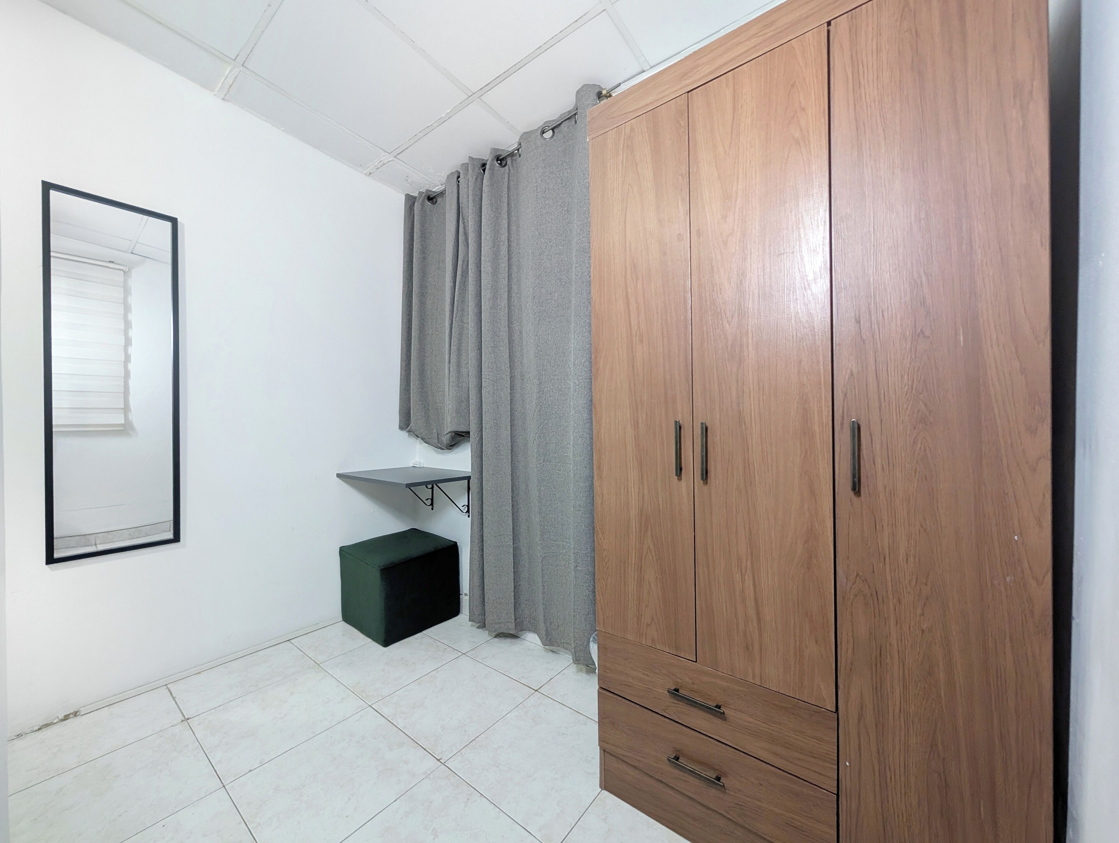 A room with a wooden wardrobe, a small green stool, a white sink, and a window with blinds.