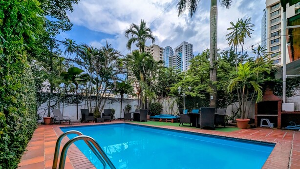 A swimming pool with a tiled deck, surrounded by greenery and outdoor furniture. - Hostal Yoha (Panama City)