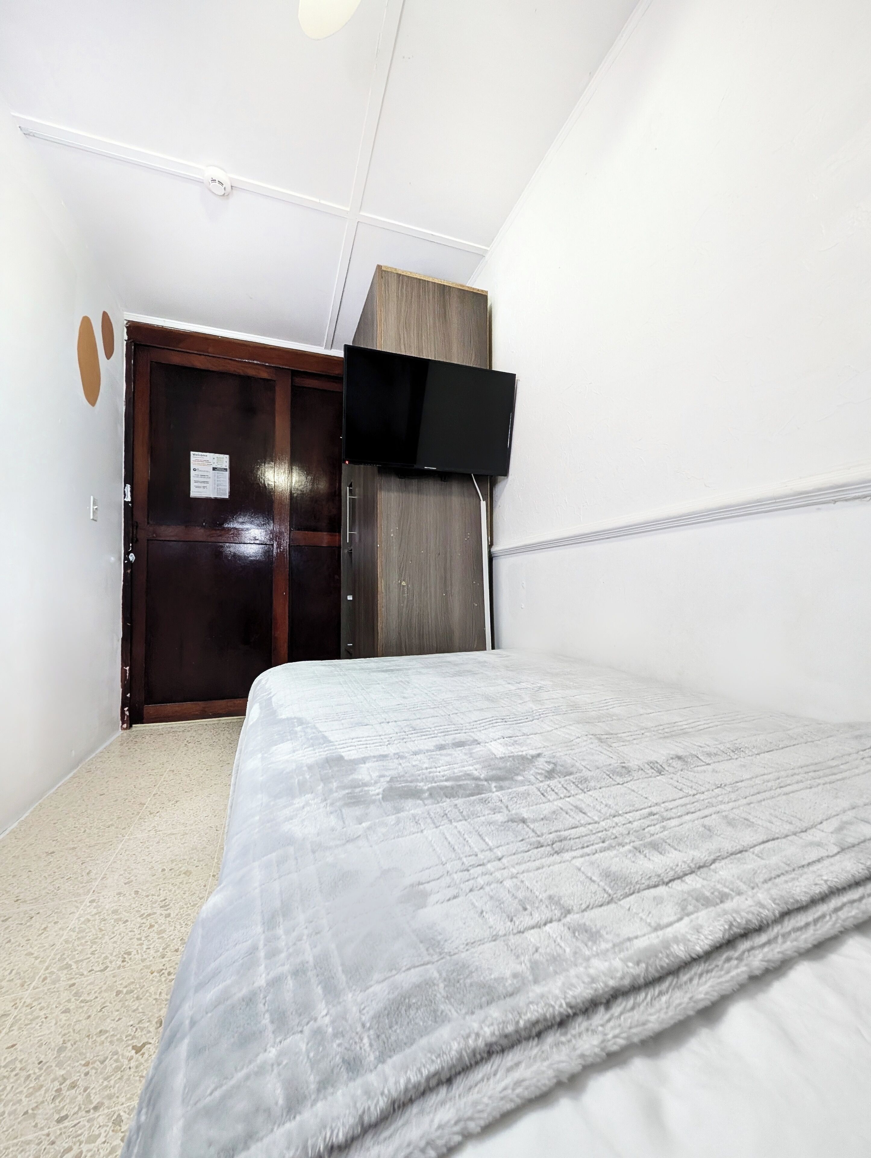 A bedroom with a bed, a wardrobe, a flat-screen TV mounted on the wall, and a ceiling light fixture.