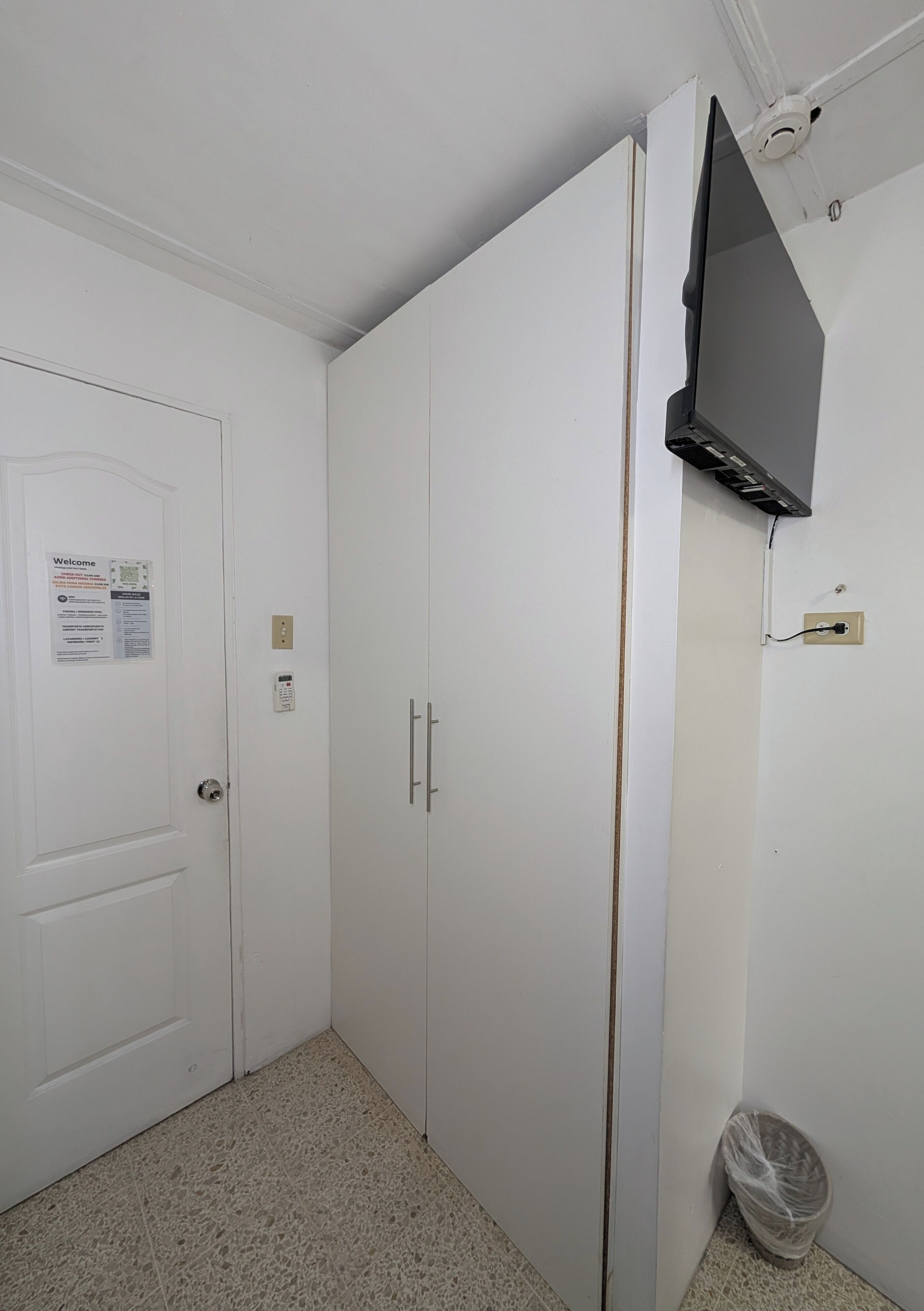 Ein Zimmer mit weißen Wänden, einer Tür mit einer Willkommensschild, einem an der Wand befestigten Flachbildfernseher und einem Mülleimer.