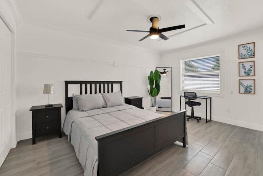 Une chambre à coucher comprenant un lit, des tables de chevet, un bureau, une chaise, un ventilateur de plafond et une fenêtre avec des stores.