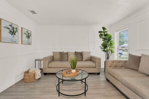 A living room with a beige sofa, a glass coffee table, and a potted plant. There are two framed artworks on the wall and a window with blinds. - #The Orlando Retreat

 (Orlando)