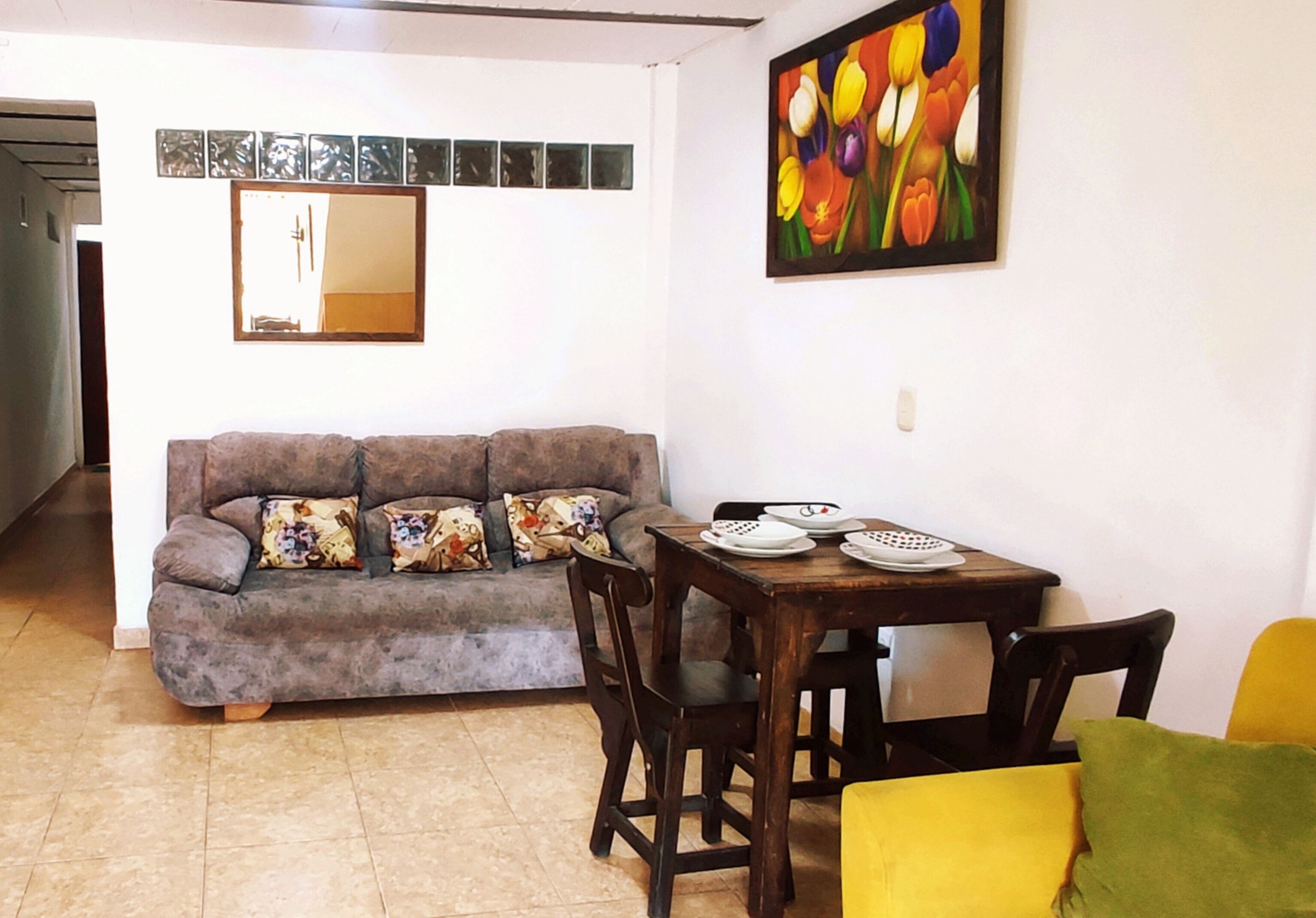 A living room with a gray sofa, a wooden dining table with chairs, and a colorful painting on the wall.