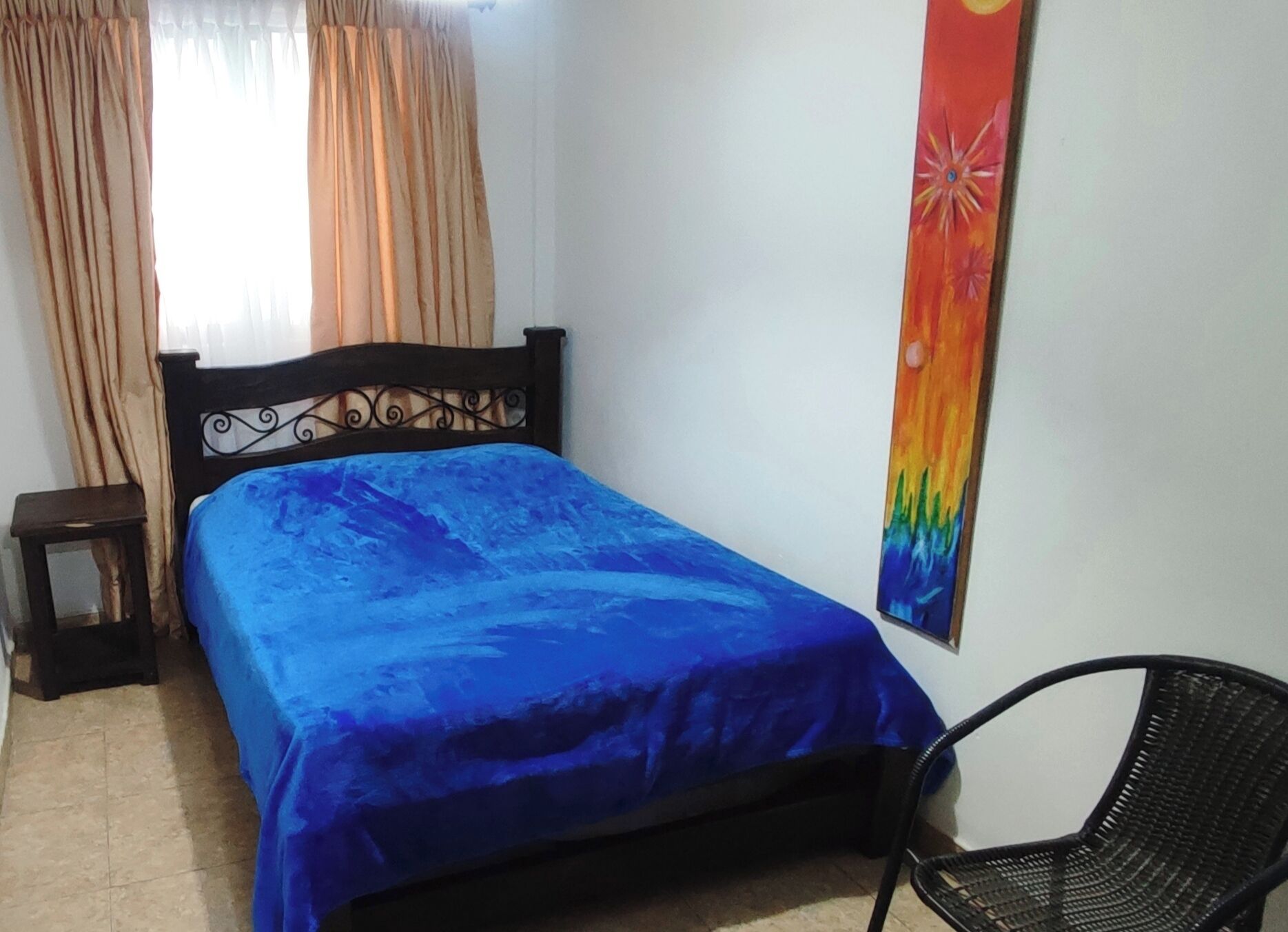 A bedroom with a bed, a chair, a small table, and a colorful wall decoration.