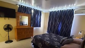 Un dormitorio con cama, cómoda, espejo, cortinas, ventilador y aire acondicionado.