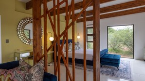 Une chambre à coucher avec un lit, un canapé bleu, une structure de plafond en bois, un grand miroir et une vue sur la verdure par une fenêtre.