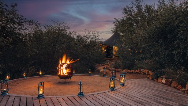 A wooden deck with a central fire feature surrounded by lanterns, set against a backdrop of trees and a twilight sky. - Canvas Villa with pool (Hoedspruit)